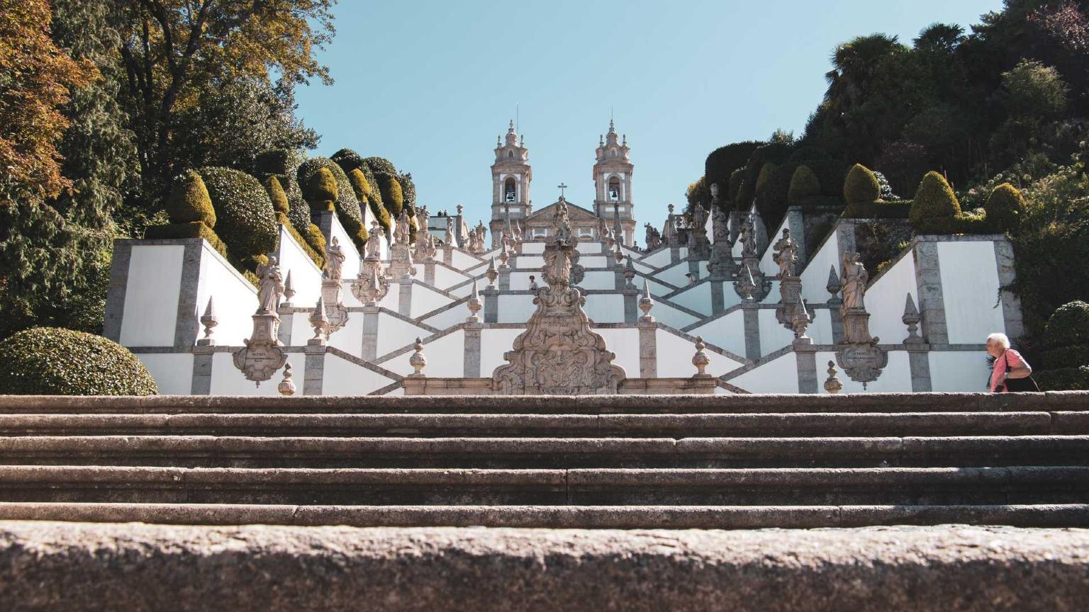 A escadaria barroca do Bom Jesus de Braga, rodeada por uma vegetação luxuriante e jardins ornamentados.