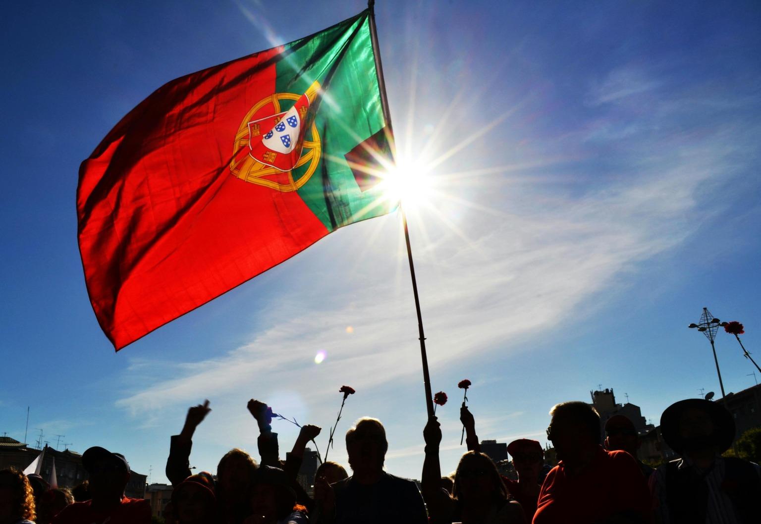 Bandeira portuguesa a tremular com pessoas a segurar cravos para celebrar a Revolução dos Cravos