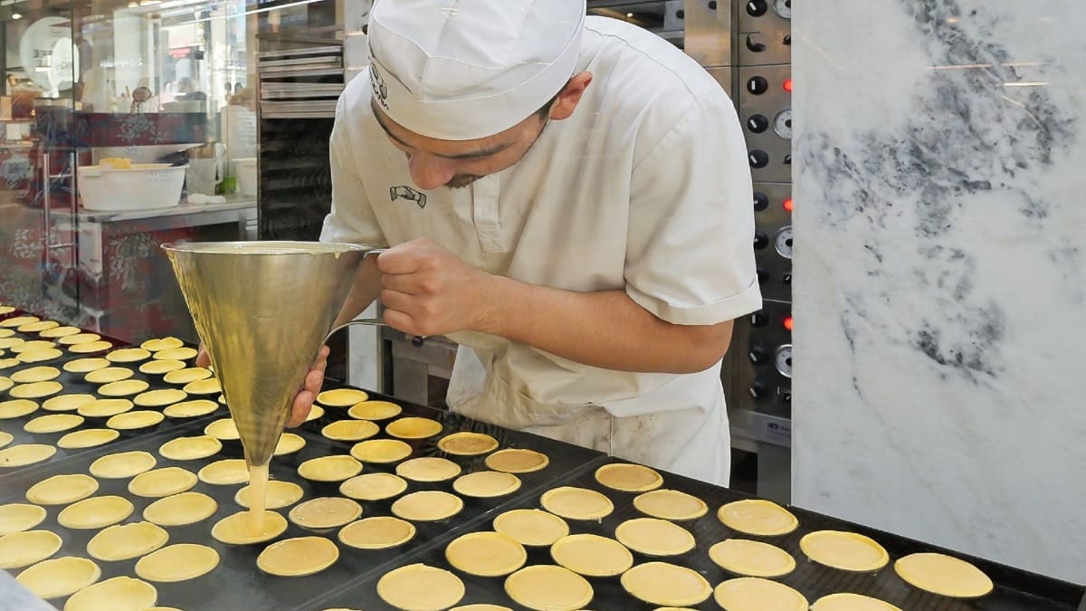 Pâtissier versant de la crème aux œufs dans des moules à pastel dans une boulangerie du centre de Porto