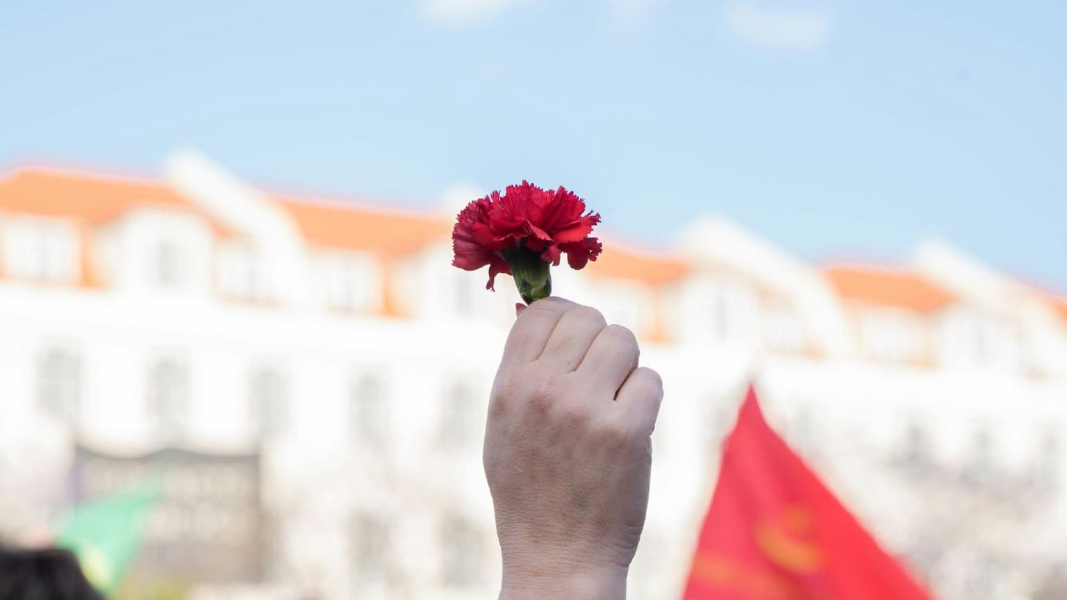 Un puño alzado sosteniendo un clavel rojo ante un cielo azul con edificios blancos y naranjas desenfocados al fondo, símbolo de la Revolución de los Claveles de 1974 y del Día de la Libertad el 25 de abril en Portugal