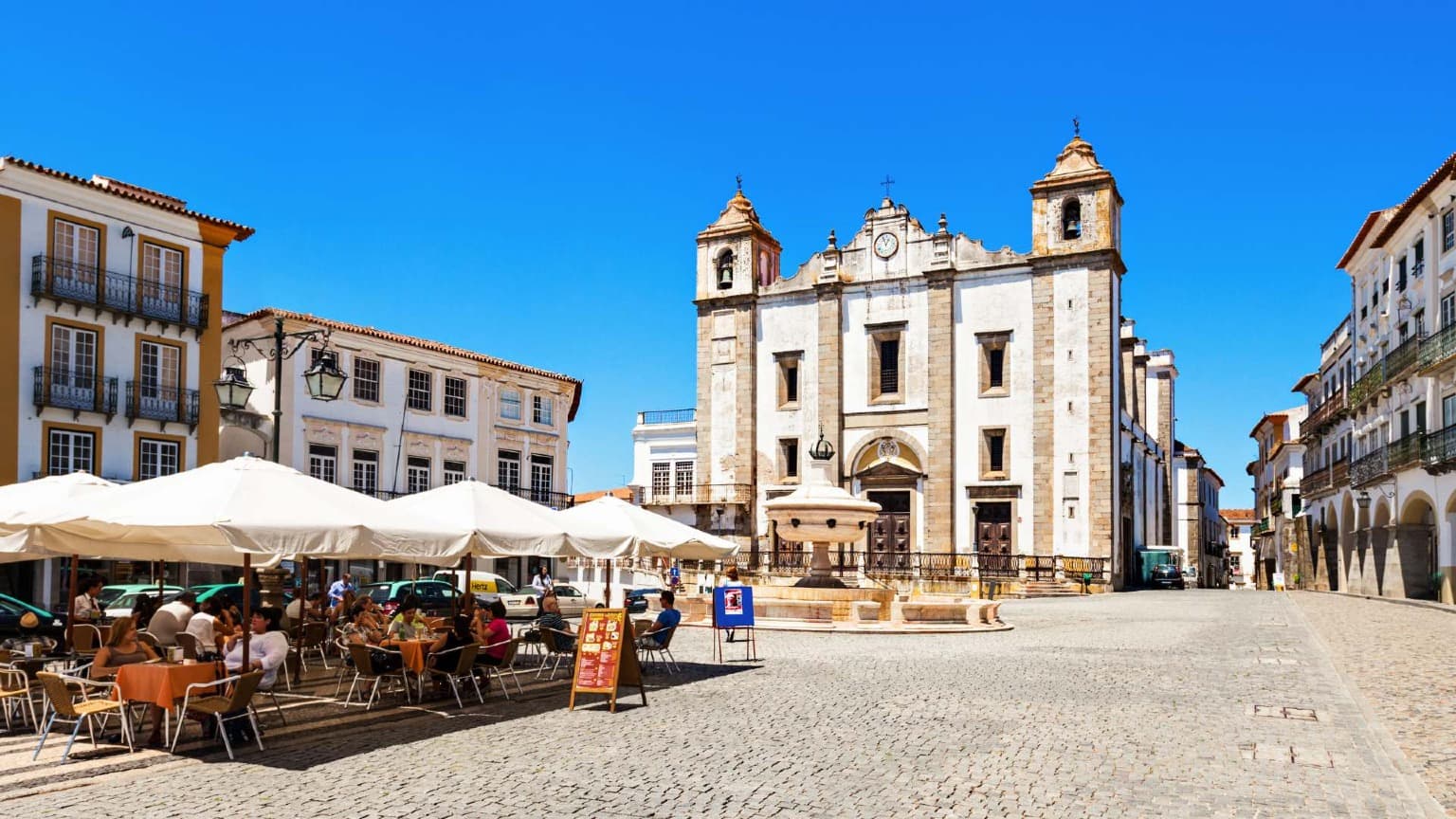 La histórica plaza principal de Évora con cafeterías al aire libre y la iglesia de Santo Antão bajo un cielo azul brillante.
