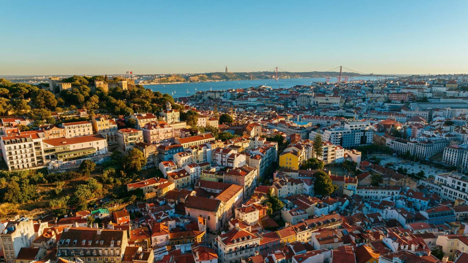 Vista aérea do centro de Lisboa mostrando o Castelo de São Jorge, o rio Tejo e a Ponte 25 de Abril ao pôr do sol.