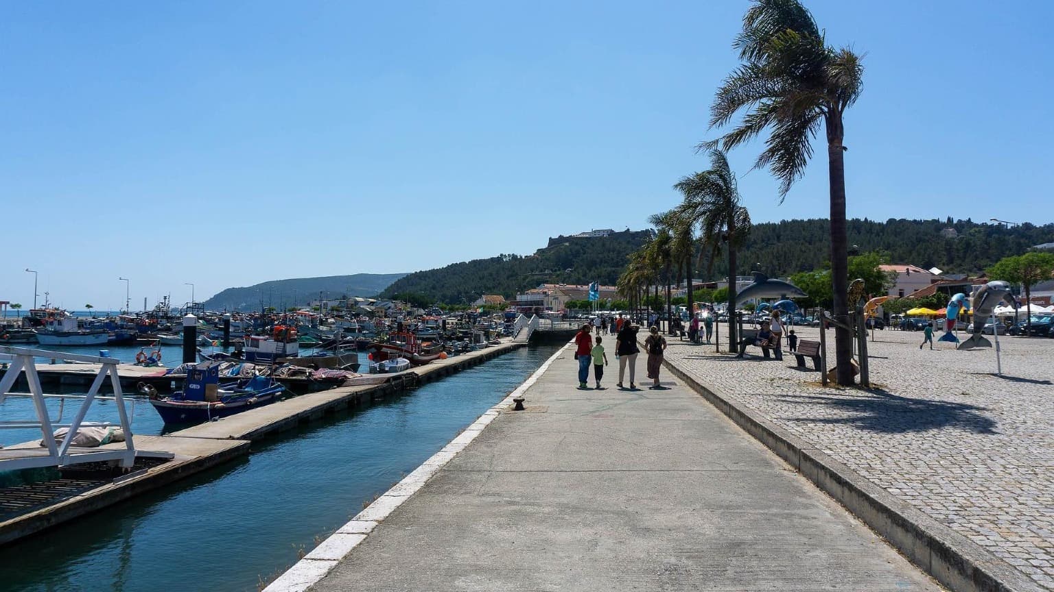 Promenade en bord de mer de l'Avenida José Mourinho à Setúbal avec des bateaux de pêche, des palmiers et une vue sur le Forte de São Filipe.