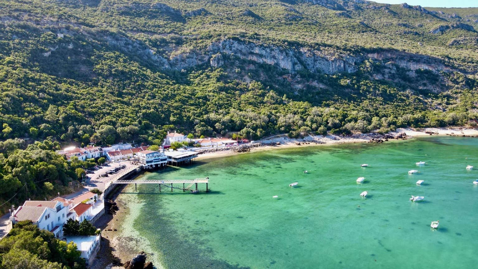 Vista aérea de Portinho da Arrábida en Setúbal con aguas esmeraldas, playa de arena y casas tradicionales junto a la ladera.
