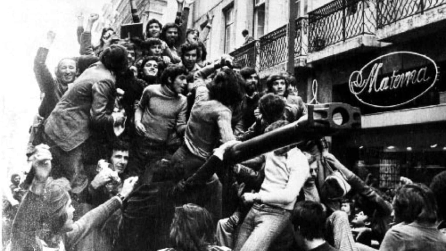 Fotografia a preto e branco de 25 de abril de 1974 mostrando multidões de civis em Lisboa a subir para viaturas militares e a celebrar a Revolução dos Cravos que pôs fim à ditadura em Portugal