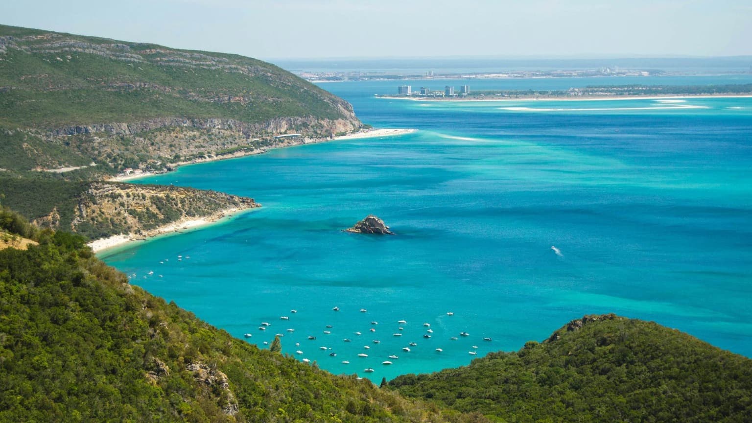 Vista panorámica del Parque Natural de Arrábida, cerca de Setúbal, con exuberantes colinas verdes, mar turquesa y barcos anclados a lo largo de la costa.