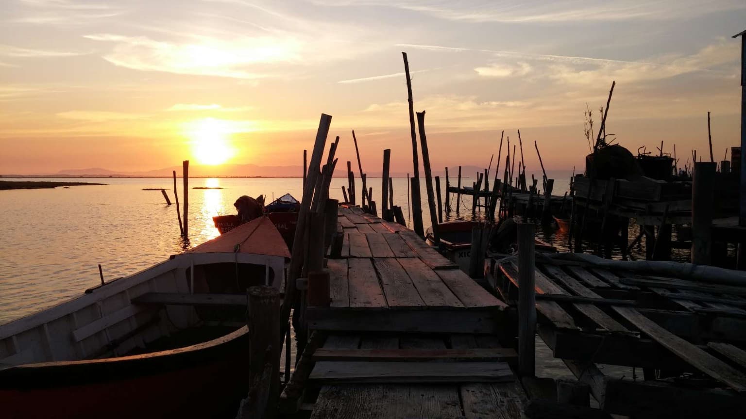 Cais palafítico de madeira da Carrasqueira ao pôr do sol com barcos de pesca tradicionais em águas calmas.