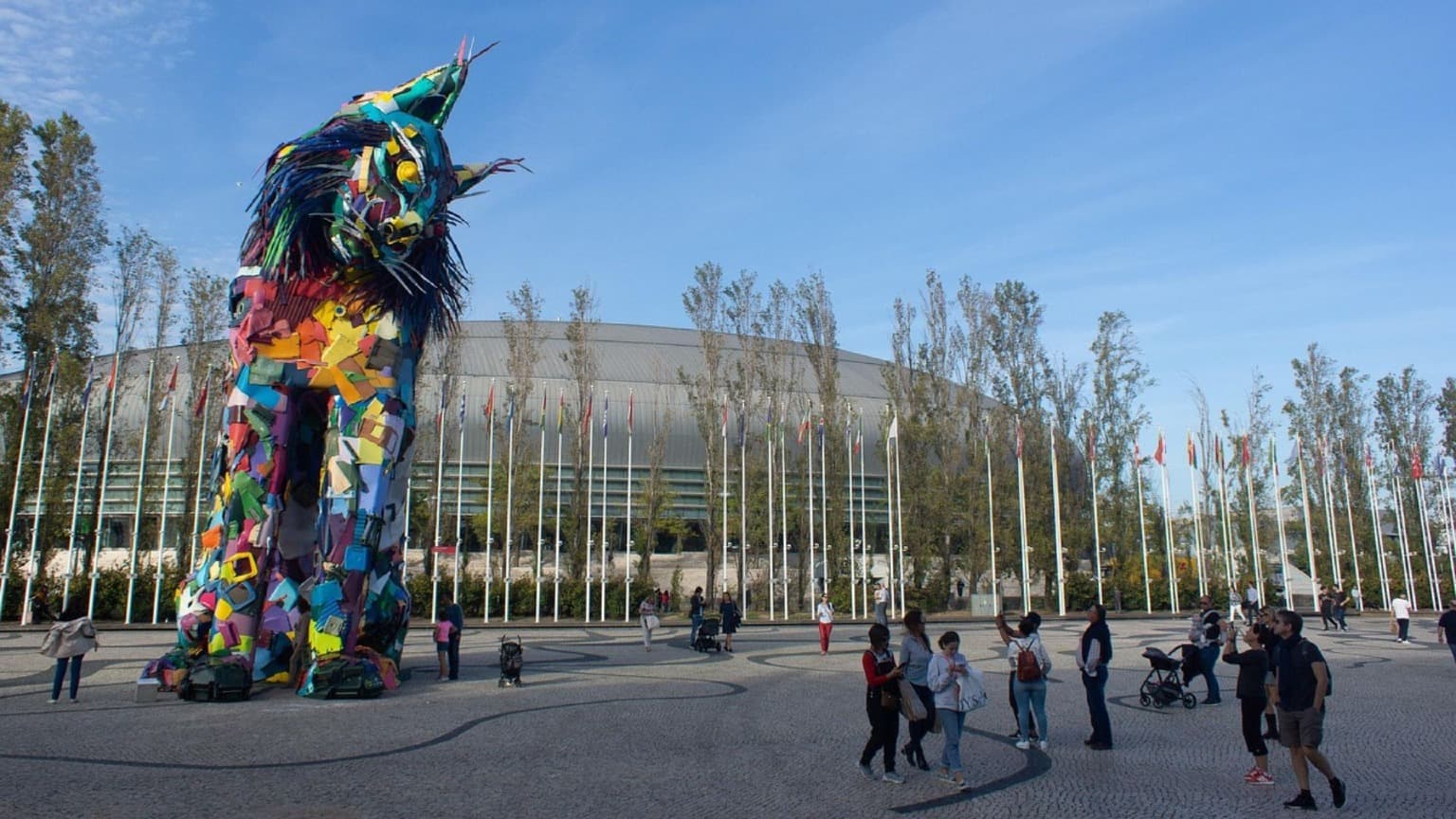 Escultura de animales de colores del artista Bordalo II realizada con materiales reciclados en el Parque das Nações de Lisboa, con banderas y el Altice Arena al fondo.