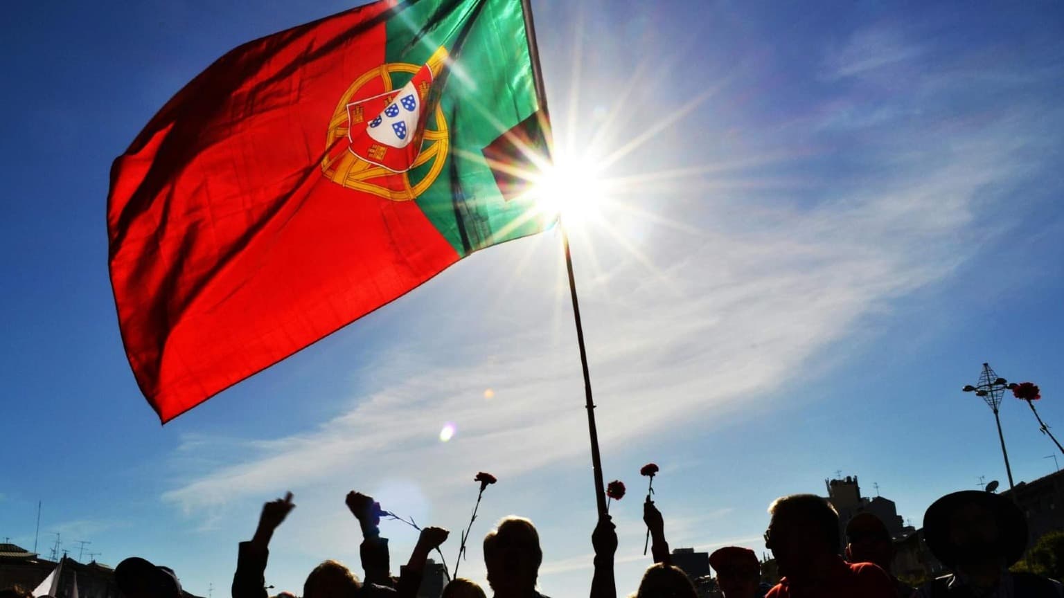 Bandeira portuguesa a tremular com pessoas a segurar cravos para celebrar a Revolução dos Cravos