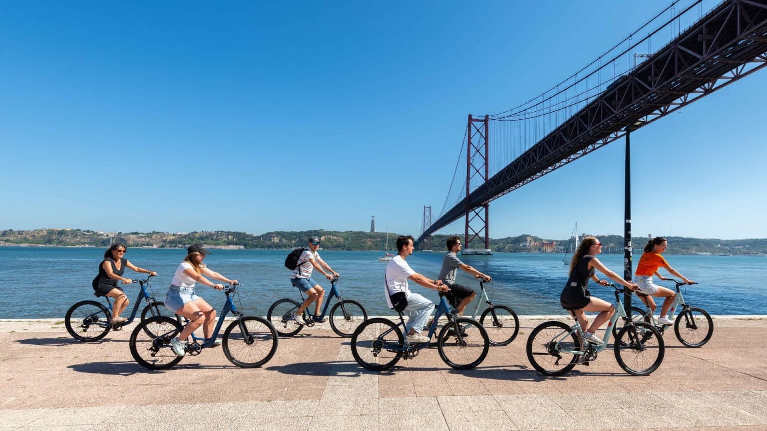 Grupo de ciclistas a pedalar ao longo do rio Tejo, sob a Ponte 25 de Abril, em Lisboa