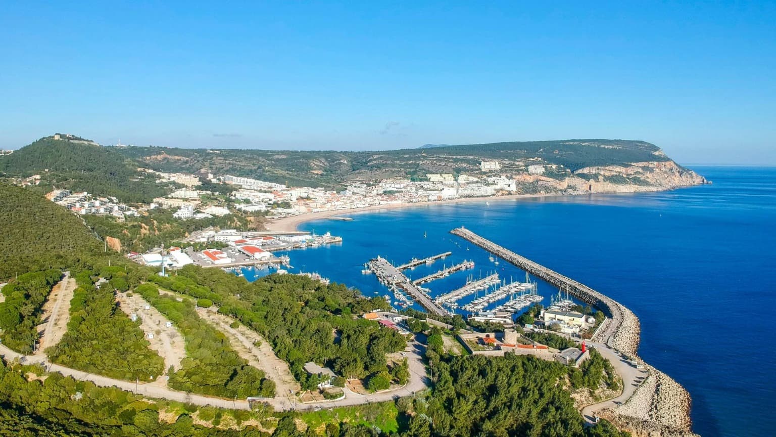 Vista aérea de Sesimbra, na região de Setúbal, com praias arenosas, porto de pesca, marina e o castelo no topo da colina.